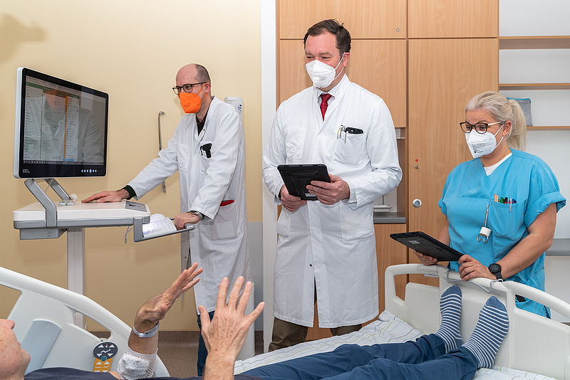 Zwei Ärzte und eine Schwester mit Mundschutz stehen an einem Patientenbett mit einem Computer und Pads in der Hand. Digitalisierung am Krankenbett sorgt für Nachhaltigkeit und mehr Zeit für Patienten.