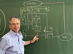 Ein Mann mit Brille und blauem Hemd steht vor einer Tafel, auf der mit Kreide bunte naturwissenschaftliche Formeln zu sehen sind