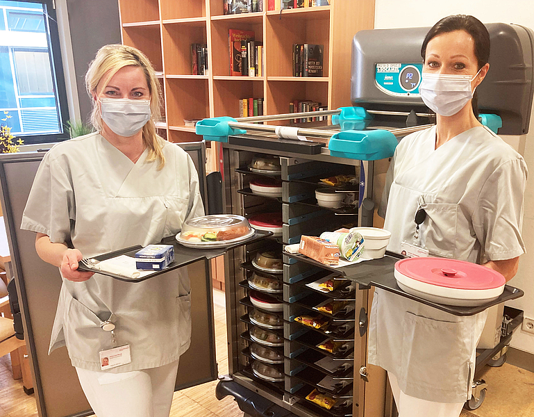 Zwei junge Krankenschwestern mit Mundschutz stehen vor einem Wagen mit Tabletts und halten je zwei Tabletts mit Essen in der Hand. Eine nachhaltige Patientenversorgung an der Unimedizin Rostock.