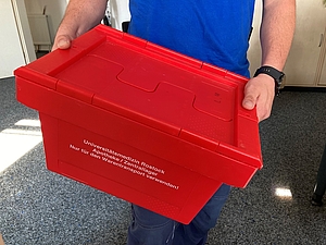 Ein roter Plastikbehälter wird von einem Mitarbeiter in einem blauen T-Shirt gehalten