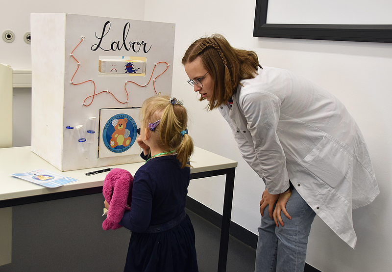 junge Frau in weißem Kittel beugt sich zu einem Mädchen mit Kuscheltier.  Beide stehen vor einem Pappkarton, der als Labor gestaltet ist. Kinder lernen den Besuch an der Unimedizin Rostock.