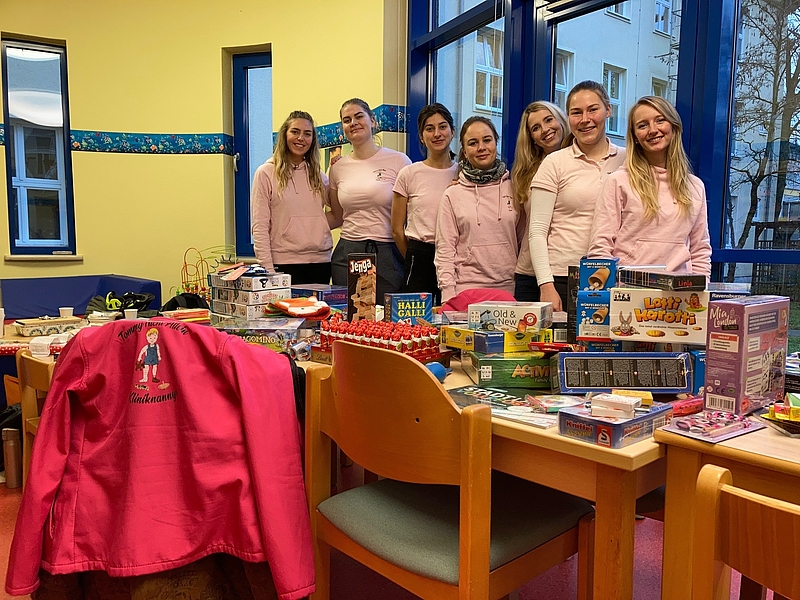 Junge Frauen in rosa T-Shirts stehen hinter einem Tisch mit Spielzeug und Süßigkeiten. Mit großer sozialer Verantwortung helfen die Kliniknannys Kindern und Eltern.