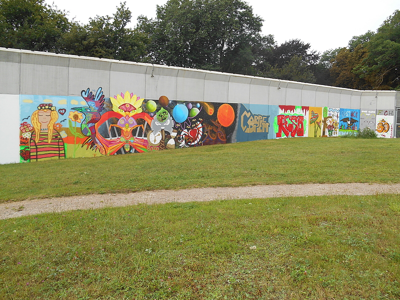 Bunte Graffitis an einer langen Mauer mit Stacheldraht. Die Universitätsmedizin Rostock setzt zahlreiche Maßnahmen im Sinne der Nachhaltigkeit um. Dazu gehören die Bereiche Umwelt, Soziales und Governance. 