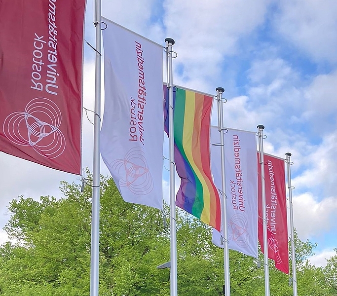 Fahnenmasten hinter blauen Glastafeln mit den Fahnen der Unimedizin Rostock und mittig der Regenbogenfahne. Das Bekenntnis zu Vielfalt gehört zur good governance.