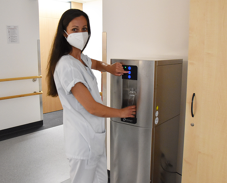 Krankenschwester mit Mundschutz benutzt einen metallfabenen Wasserspender auf einem Flur. Nachhaltigkeit an der Unimedizin Rostock betrifft auch die Versorgung der Patienten mit leitungsgebundenen Trinkwasser.