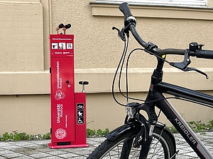 Rote Säule mit Fahrradzubehör und Fahrrad vor einer Hauswand.