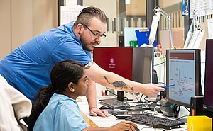 Pflger mit Brille und Bart erklärt einer sitzenden indischen Pflegerin etwas am Computer.