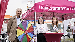 3 Personen schauen freundlich nach vorne, vor ihnen steht ein Glücksrad, 2 von ihnen stehen unter einem roten Pavillion.