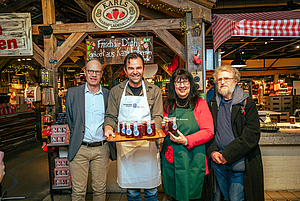 Vier Personen, zwei davon mit Kochschürzen, präsentieren ein Tablett mit Fruchtaufstrichgläsern
