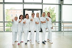 Sieben Frauen in weißer Dienstkleidung stehen in einem Krankenhausflur und machen die Geste der Zustimmung "Daumen hoch".
