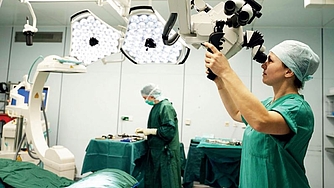 Zwei medizinische Fachkräfte in Operationskleidung in einem Operationssaal, eine bedient ein großes Operationsmikroskop, die andere steht am Instrumententisch.