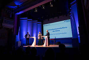 Drei Personen stehen auf einer Bühne mit zwei weißen Stehtischen vor einer Leinwand mit der Projektion 'Nachwuchswissenschaftlerin des Jahres Professorin Dr. Anna Lene Seidler'