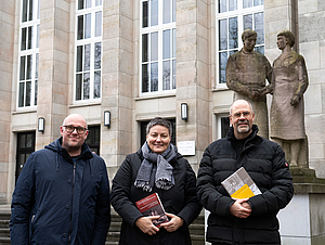 Die Arbeitsgruppe vom Arbeitsbereich Geschichte der Medizin steht vor einem Hörsaal-Gebäude im Bauhausstil. Im Hintergrund sind DDR-Skulpturen zu sehen