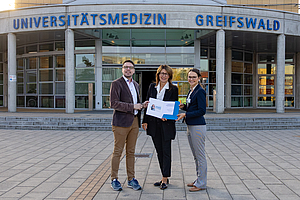 3 Personen stehen nebeneinander, halten einen Hefter in der Hand un grinsen. Sie stehen vor der Universitätsmedizin Greifswald.