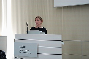 Person steht hinter einem Rednerpult mit dem Schild 'Pommersches Landesmuseum' und spricht in ein Mikrofon