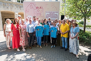 Gruppe von Menschen steht vor einem großen Schild mit Text und Logo im Freien an einem sonnigen Tag.
