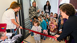Eine Gruppe Kinder schneidet ein rotes Band im Vordergrund durch. Links und rechts stehen Erwachsene