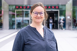 Nahaufnahme einer Frau mit zusammengebundenen dunklen Haaren und Brille vor einem Gebäudeeingang
