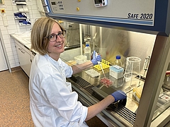 Eine Frau im weißen Kittel sitzt an einer Laborbank und hält eine Pipette in der Hand.