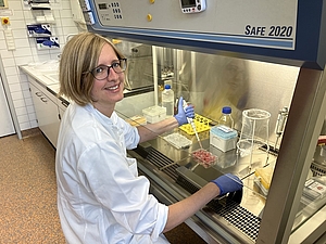 Eine Frau im weißen Kittel sitzt an einer Laborbank und hält eine Pipette in der Hand.