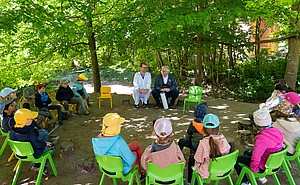 Kinder sitzen im Kreis unter Bäumen und hören zwei Männern zu