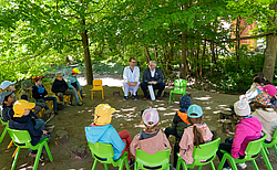 Kinder sitzen im Kreis unter Bäumen und hören zwei Männern zu