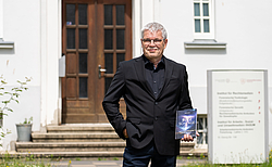 älterer Mann mit dunkler Kleidung hält Buch in der Hand und steht vor dunkler Tür