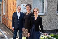 Drei Personen stehen nebeneinander vor einem Gebäude, zwei Frauen und ein Mann.