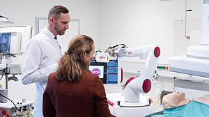 Eine Person im weißen Kittel zeigt einer anderen Person einen Roboter.