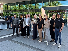Eine Gruppe von elf Personen posiert lächelnd vor dem Haupteingang der Universitätsmedizin Rostock für ein gemeinsames Foto auf dem gepflasterten Vorplatz.