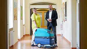 Ein Mann und eine Frau stehen in einem hellen Flur, vor ihnen steht eine große Maschine zur Reinigung
