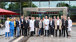 Gruppenfoto mit vielen Menschen, teilweise im Arztkittel und Pflegekasack, vor dem modernen Haupteingang der Unimedizin