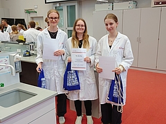 Drei Studentinnen in weißen Kitteln stehen in einem Labor und halten Beutel und Urkunden in den Händen. Im Hintergrund sind weitere Studenten zu sehen.
