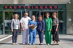 Sechs Personen stehen vor einem Gebäude mit der Aufschrift 'HAUPTEINGANG', darunter zwei in medizinischer Kleidung und eine mit einem Blumenstrauß
