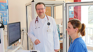 Arzt mit Kittel und Stethokop neben einer Pflegekraft im blauen Kasack in einem Untersuchungsraum