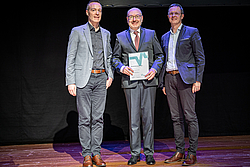 Drei Männer in Anzügen stehen auf einer Bühne, der mittlere hält eine Auszeichnung in der Hand
