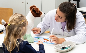 Ein Kind von hinten und eine Studentin ihr gegen über am Tisch sitzend untersucht ein Kuscheltierhund