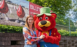 Frau in bunter Bluse übergibt rotes Kuschelkissen an eine Erdbeerfigur mit Hut