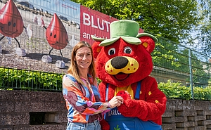 Frau in bunter Bluse übergibt rotes Kuschelkissen an eine Erdbeerfigur mit Hut