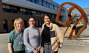 Drei junge Frauen stehen vor einem großen Gebäude, im Hintergrund ist ein großes Kunstobjekt