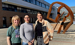 Drei junge Frauen stehen vor einem großen Gebäude, im Hintergrund ist ein großes Kunstobjekt
