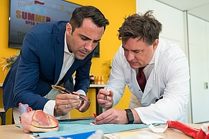 Zwei Männer in Anzug und weißem Kittel arbeiten konzentriert an einem Modell auf einem Tisch mit medizinischen Requisiten