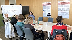 Mehrere Personen sitzen an einem Konferenztisch in einem Schulungsraum mit Präsentationsfolie und Informationsplakaten an der Wand