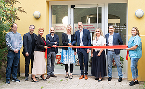 Neun Personen stehen vor einem Eingang, der mit kindgerechten Motiven und dem Schriftzug Traumaambulanz beschriftet ist, und schneiden ein rotes Band durch