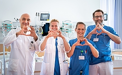 Zwei Männer und zwei Frauen formen mit den Händen Herzen. Im Hintergrund stehen medizinische Geräte.