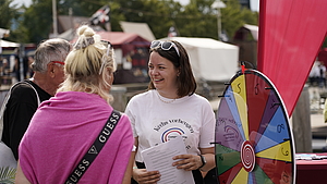 Eine lächelnde Person neben einem Glücksrad schaut zu 2 weiteren Personen