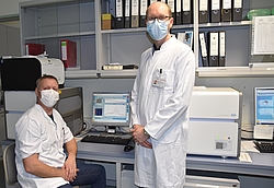 Dr. Köller und Dr. Warnke in weißen Kitteln in einem Büroraum vor einem Computermonitor
