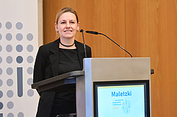 Frau steht lächelnd hinter einem Rednerpult mit Mikrofonen und einem Bildschirm, auf dem 'Maletzki' zu lesen ist