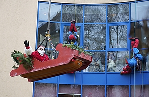 Der Nikolaus sitzt in einem Schlitten und winkt. Er hängt vor einem Gebäude.