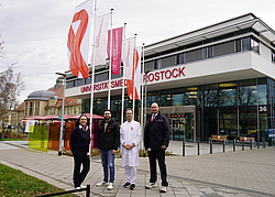 Vier Personen stehen vor dem Haupteingang. Hinter ihnen wehen die Flaggen der Unimedizin und die Welt-Aids-Flagge (eine weiße Flagge mit roter Schleife)